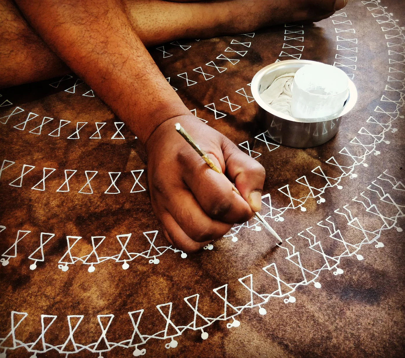 A close-up of a person's hand drawing white warli patterns on a brown textured surface, with a small metal bowl of white pigment made of rice nearby.
